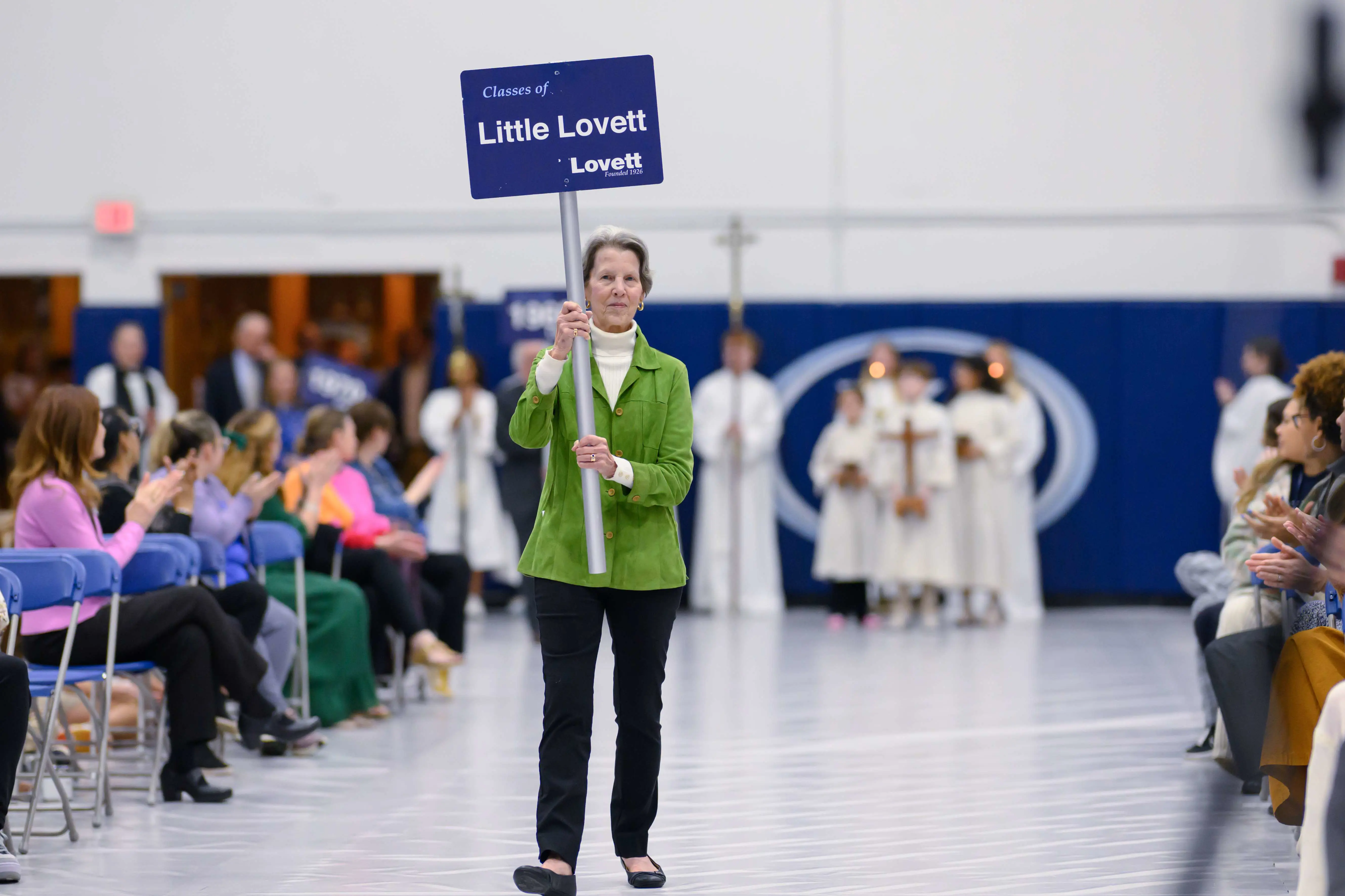 A photograph of Fontaine Draper, representing the classes of Little Lovett, smiling as she walks across a gymnasium floor holding up a blue sign that reads 'Classes of Little Lovett.'